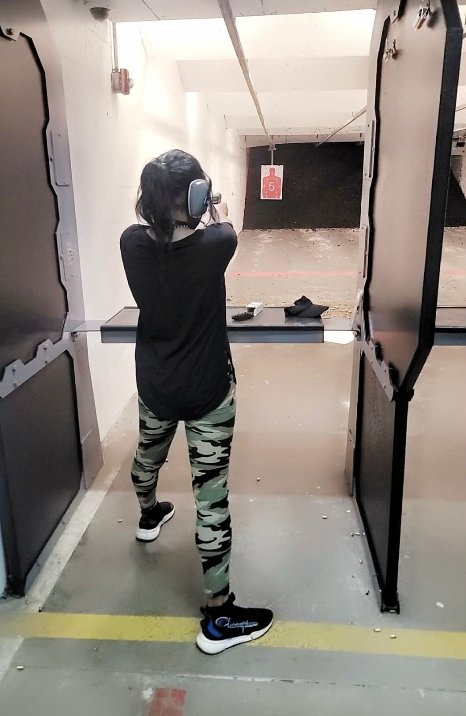 Person wearing headphones and camouflage pants aiming a gun at a shooting range target indoors