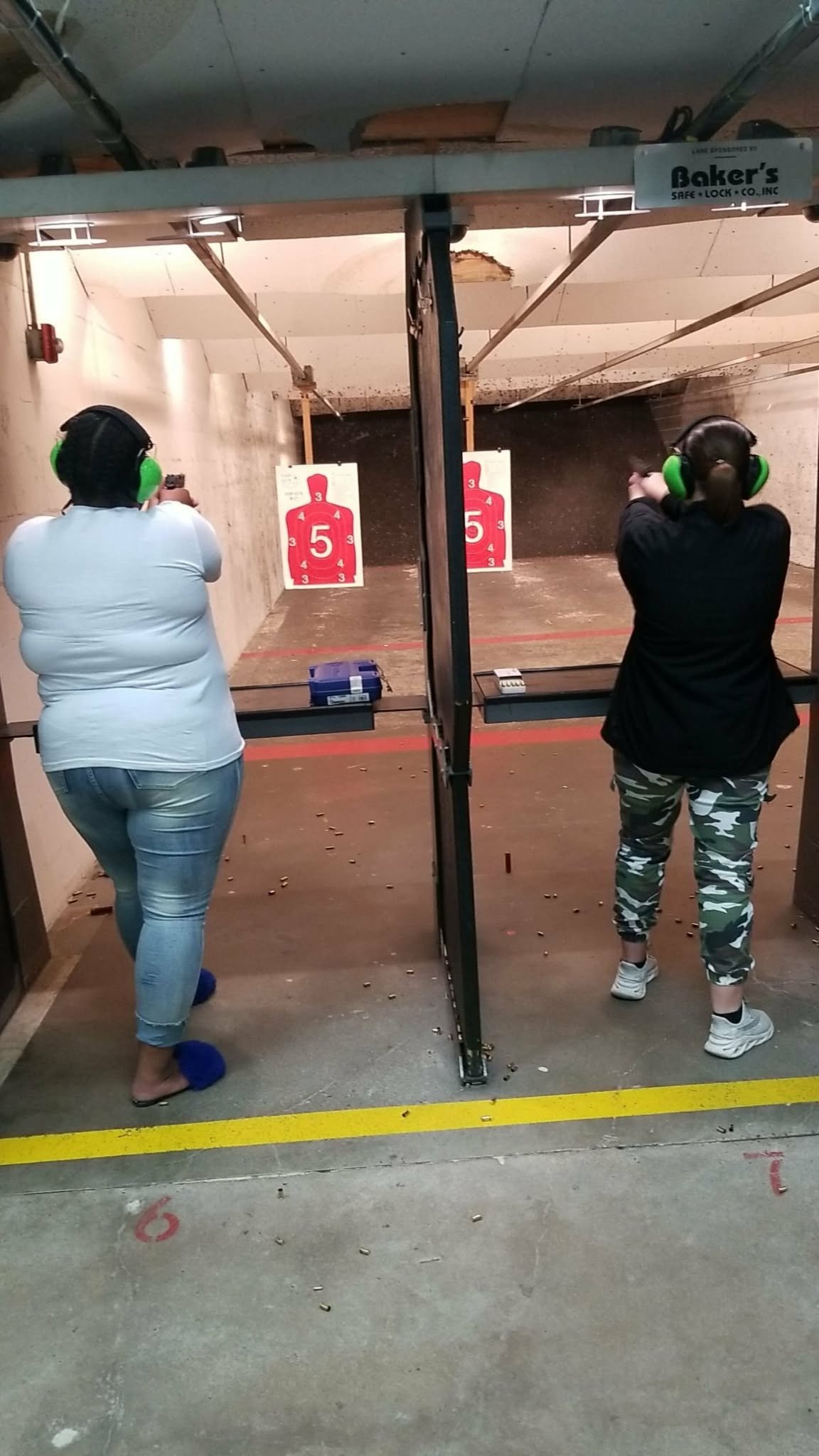 Two shooters wearing hearing protection at an indoor gun range, aiming at red and white target silhouettes downrange.