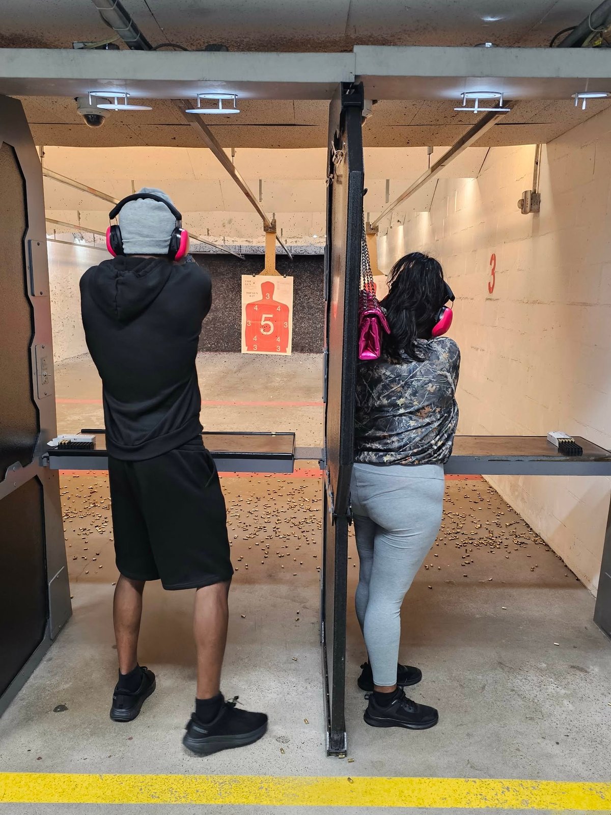 Two people wearing protective gear at indoor shooting range, aiming at paper targets downrange
