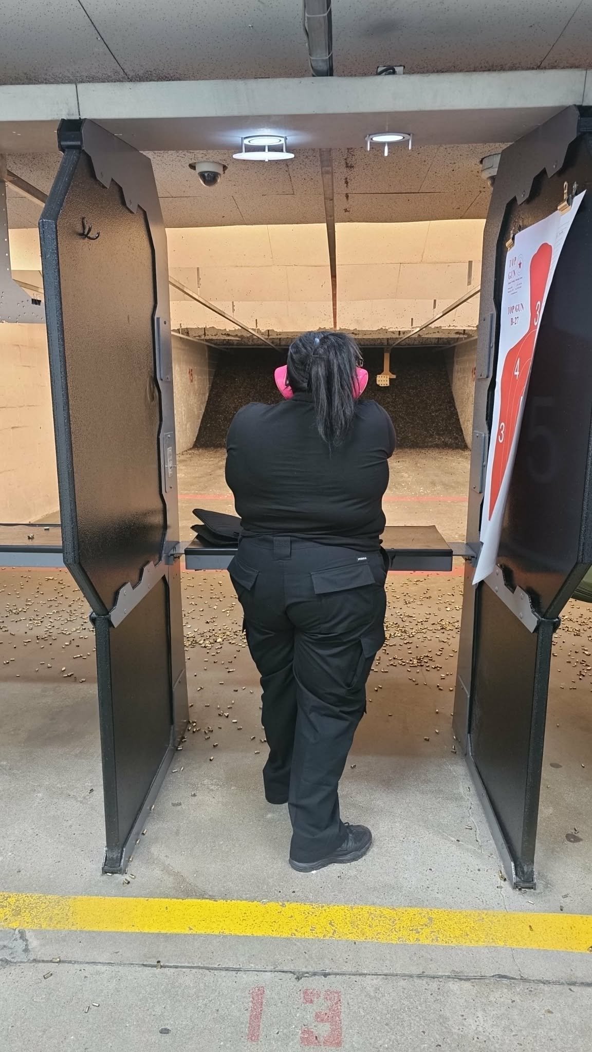 Person in black outfit and pink ear protection shooting at target in indoor shooting range stall