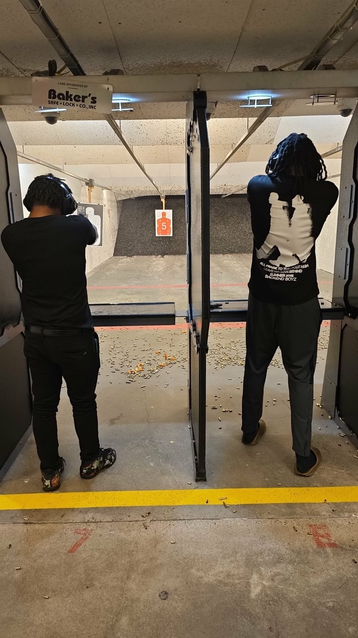 Two people in black clothing at an indoor shooting range, standing at separate shooting stations with targets visible downrange