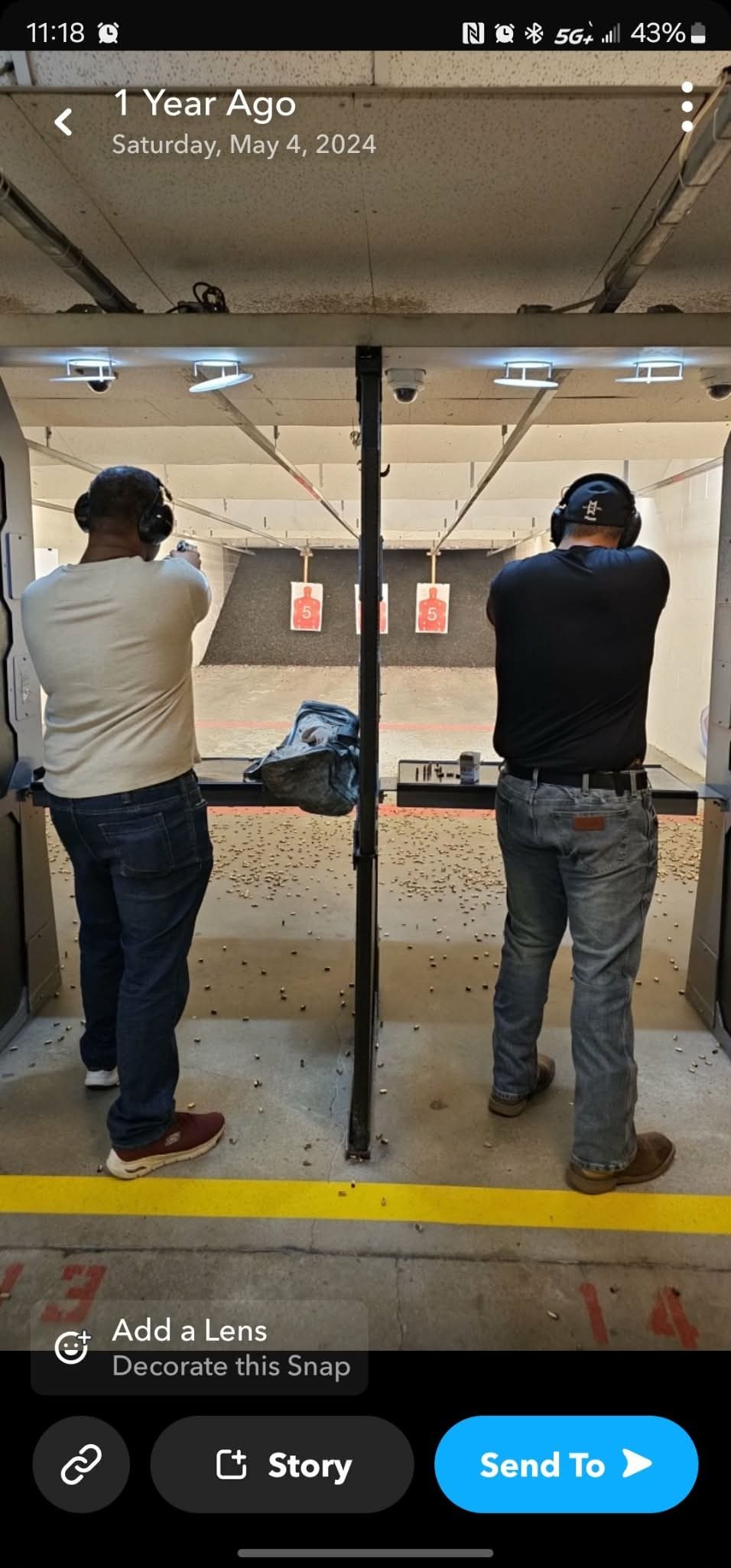 Two people wearing protective headgear at an indoor shooting range, aiming at targets from separate stalls separated by a divider