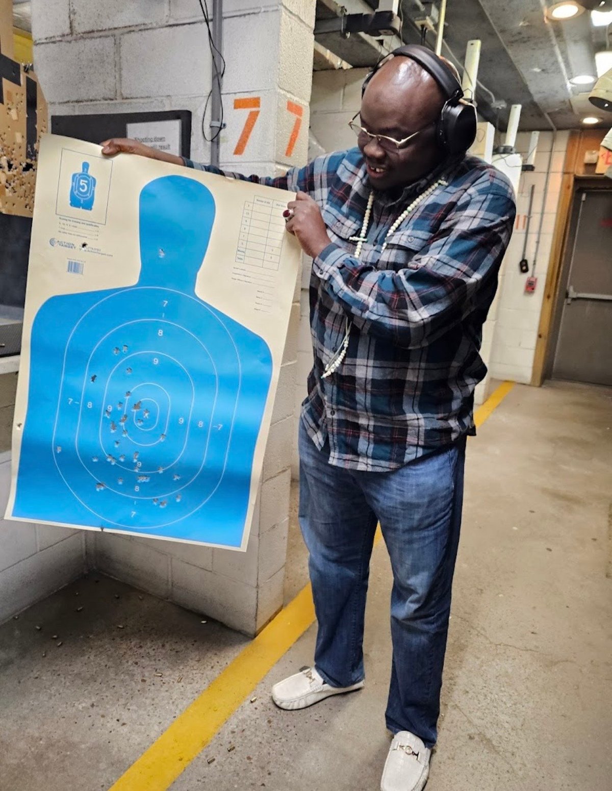 Man wearing headphones and plaid shirt inspecting a blue target at an indoor shooting range