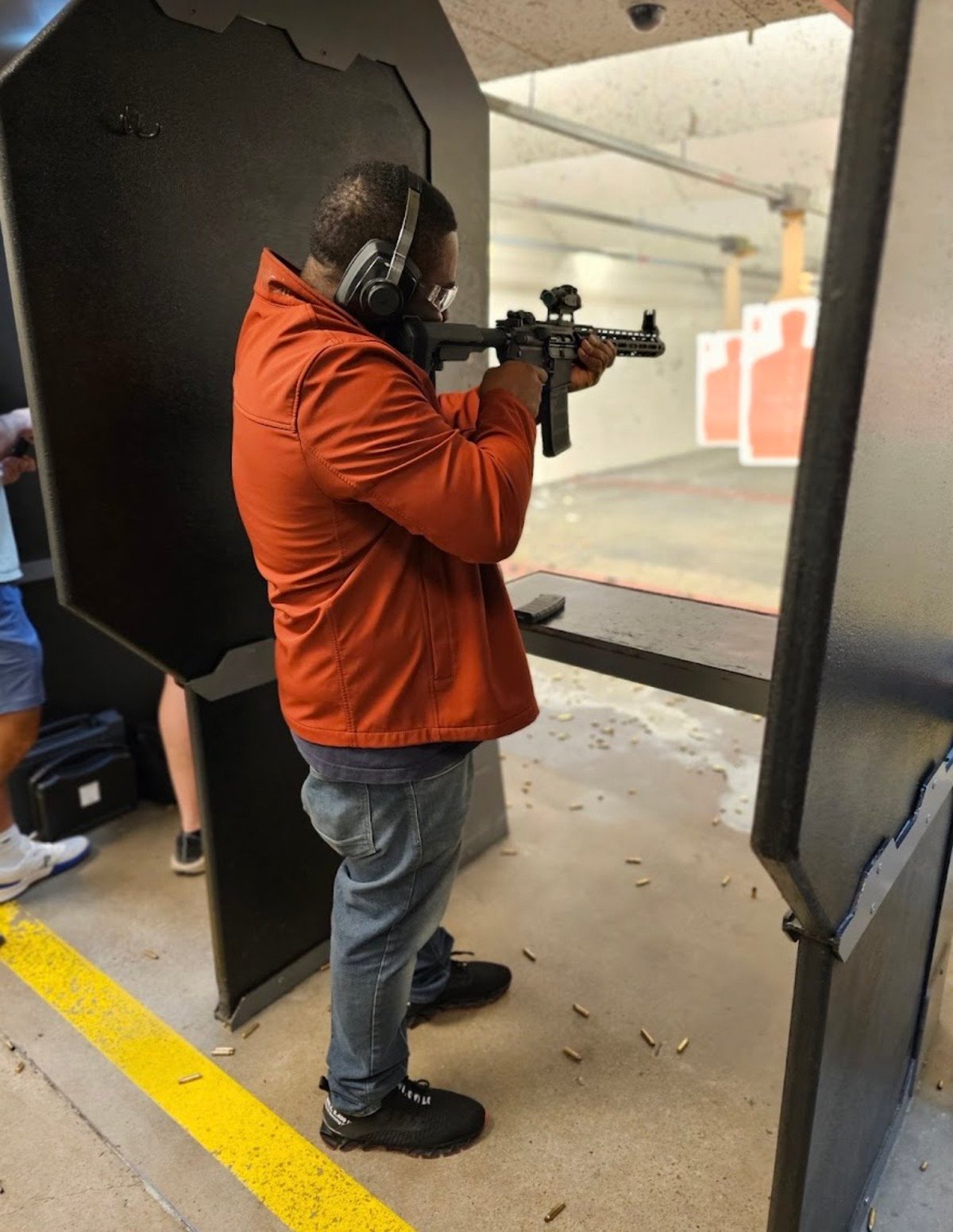 Person in orange jacket aiming rifle at target at indoor shooting range with protective barrier