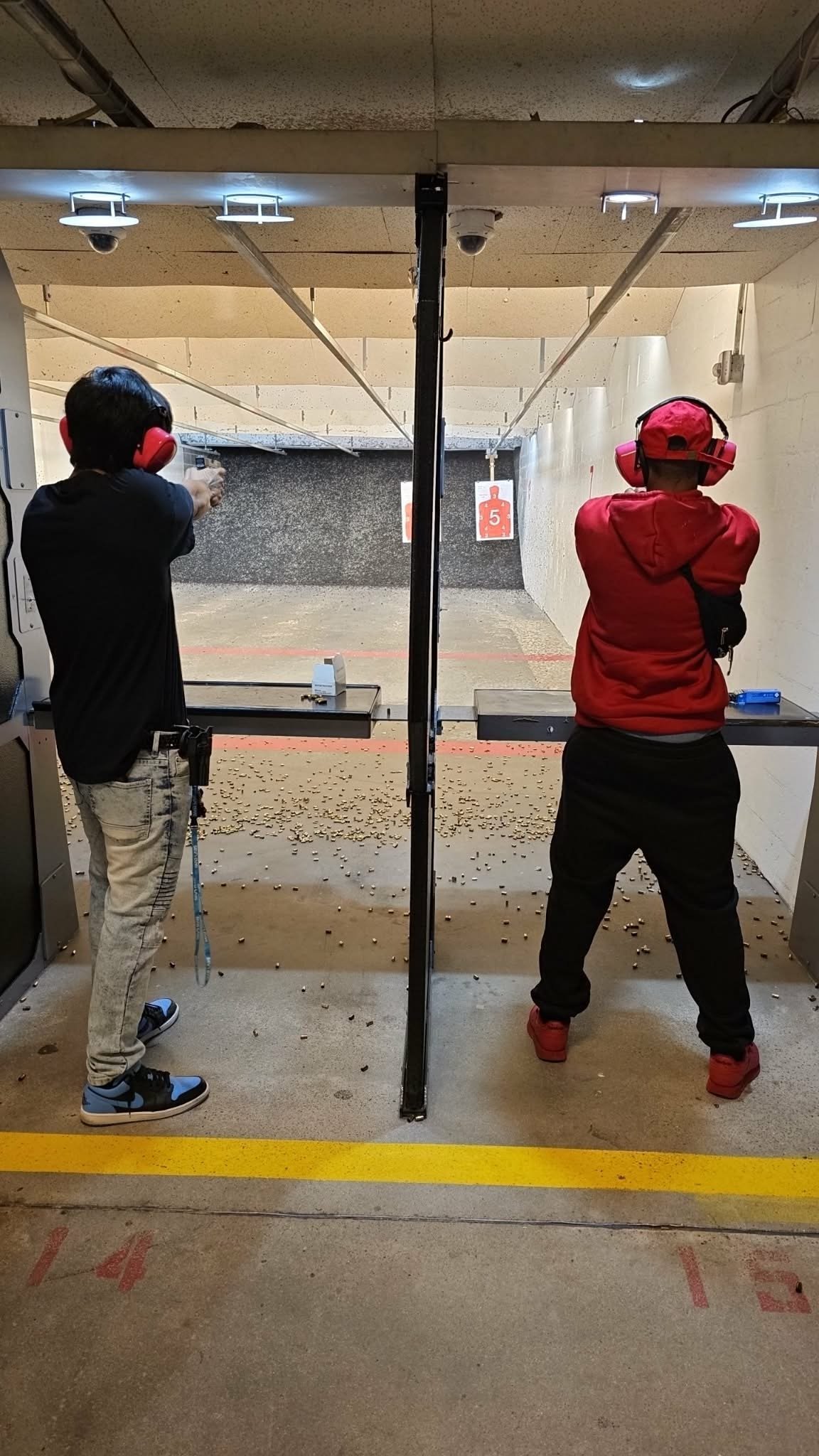 Two shooters at an indoor gun range, one in black and one in red, aiming at targets downrange