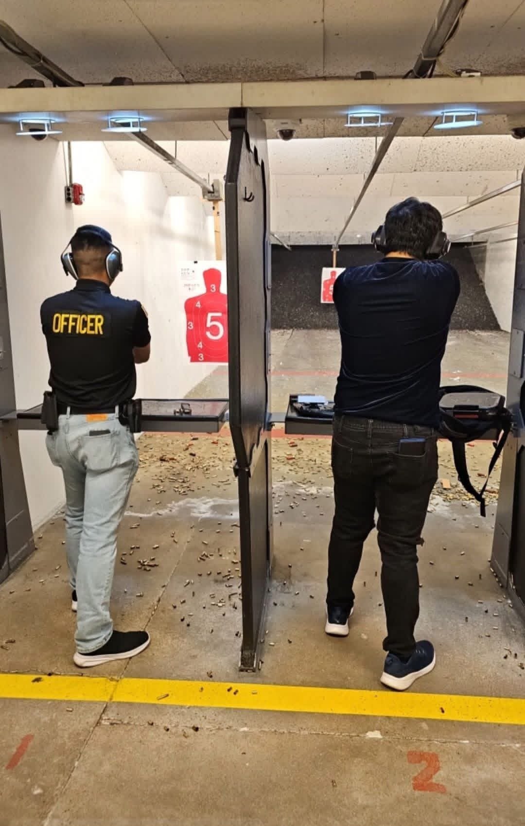 Two people in separate shooting lanes at an indoor range, one wearing an Officer shirt, aiming at red target papers downrange