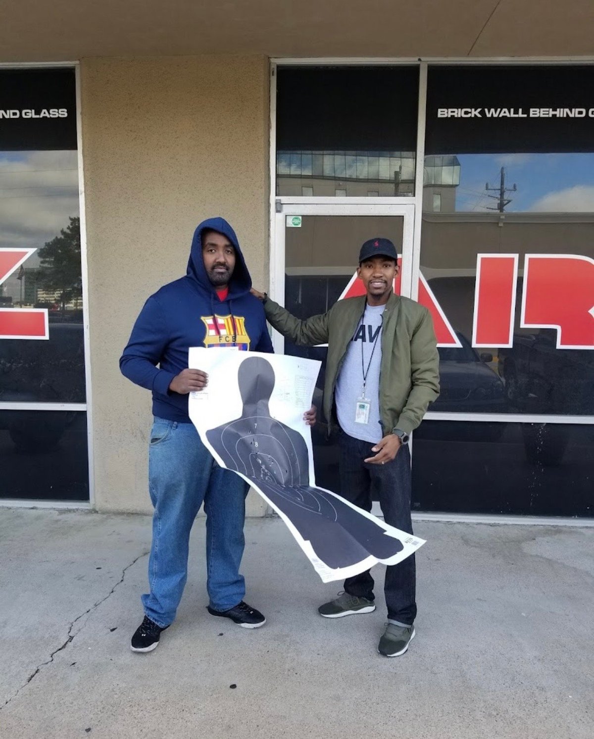 Two men hold a target shooting cutout in front of a building with signage and glass storefront