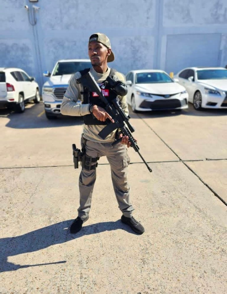 Person in tan cap and tactical gear holding rifle in parking lot with white vehicles in background