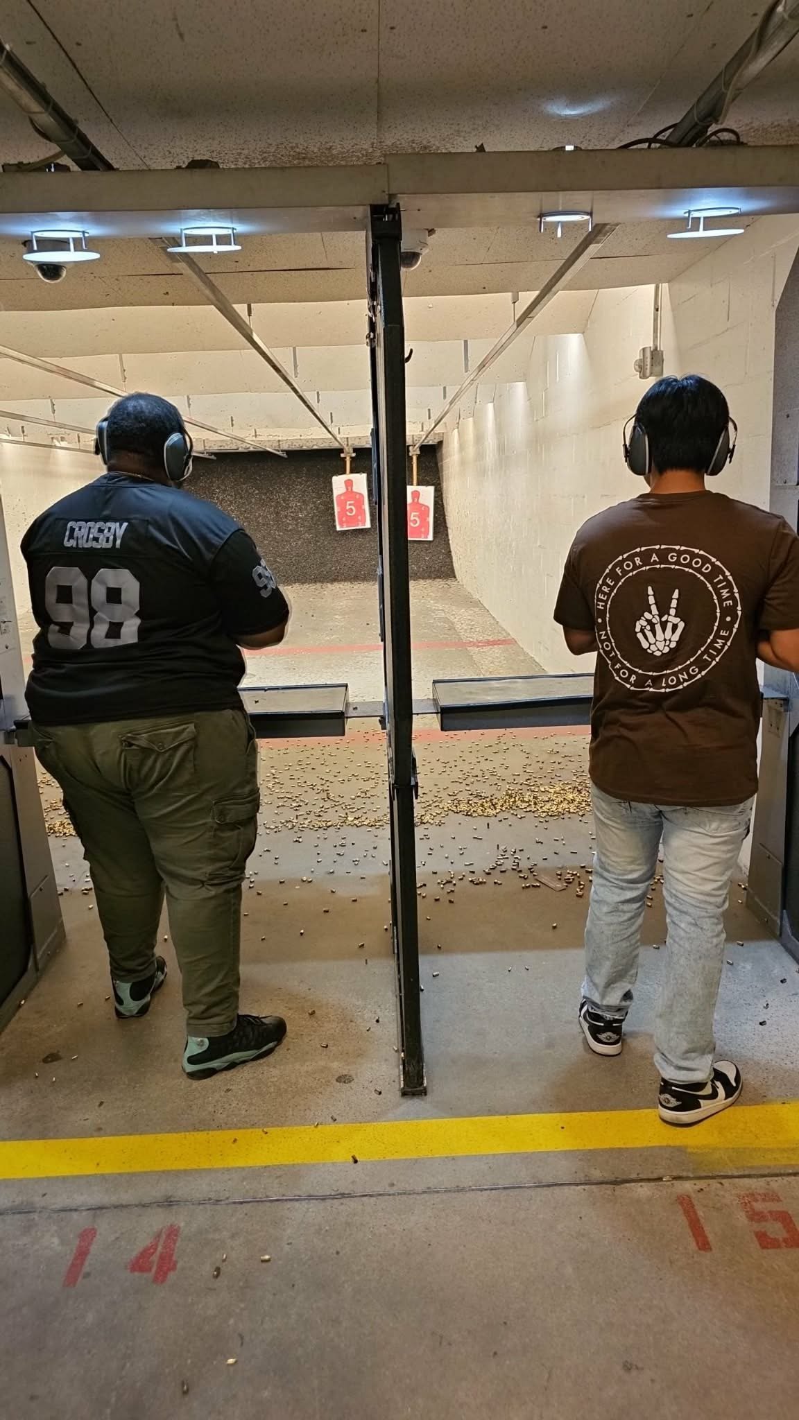 Two people wearing headphones at a shooting range, standing at separate stalls with targets ahead