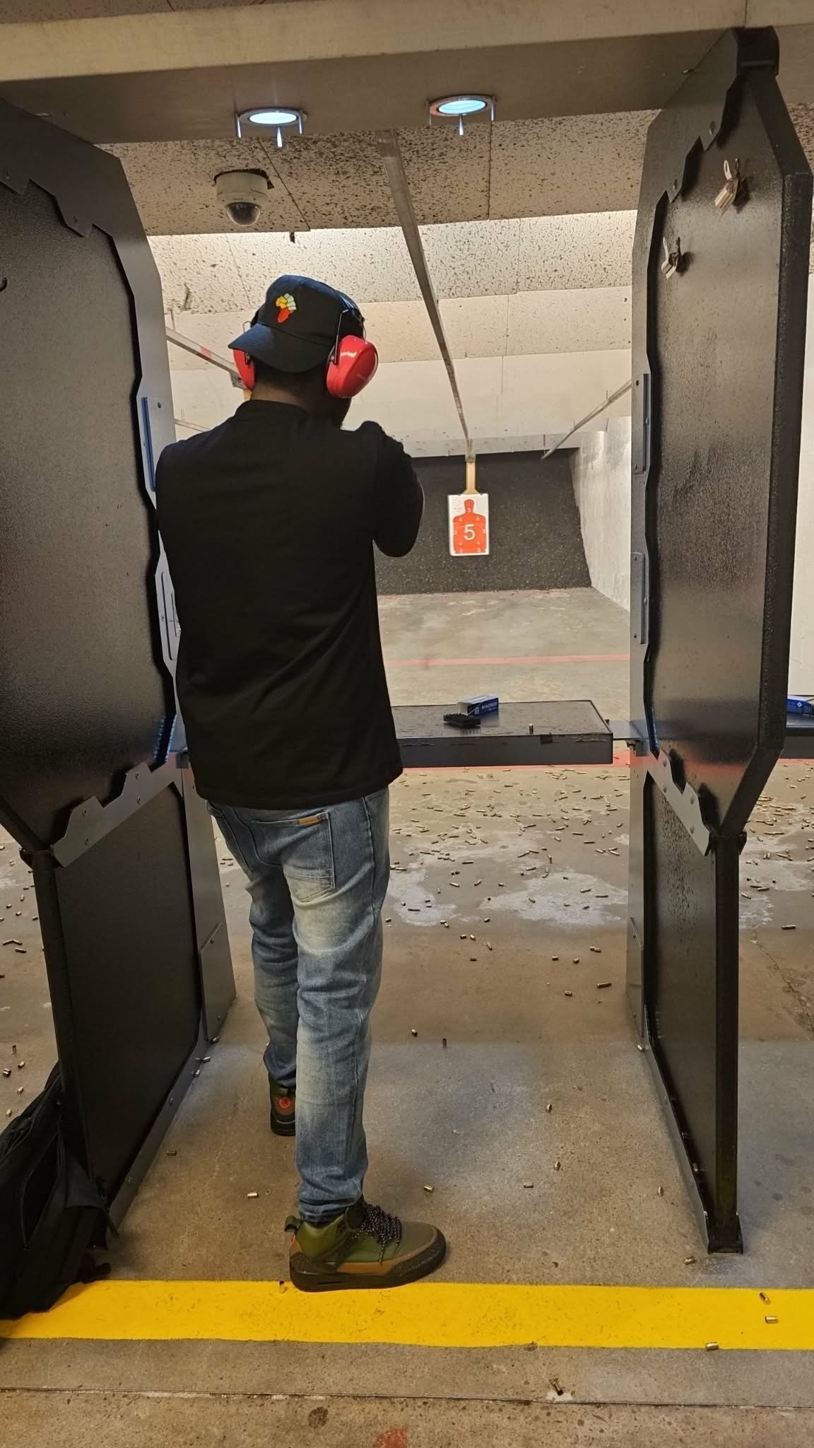 Person at indoor shooting range wearing red earmuffs and black jacket aiming at target downrange