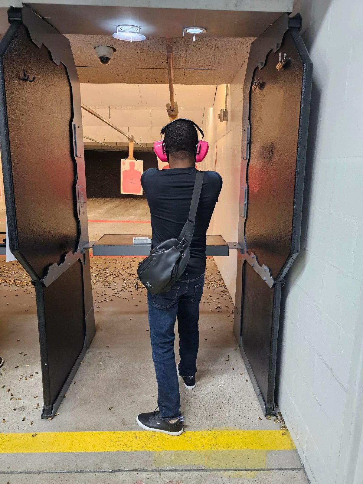 Person wearing pink headphones and black shoulder bag entering an indoor shooting range, viewed from behind.