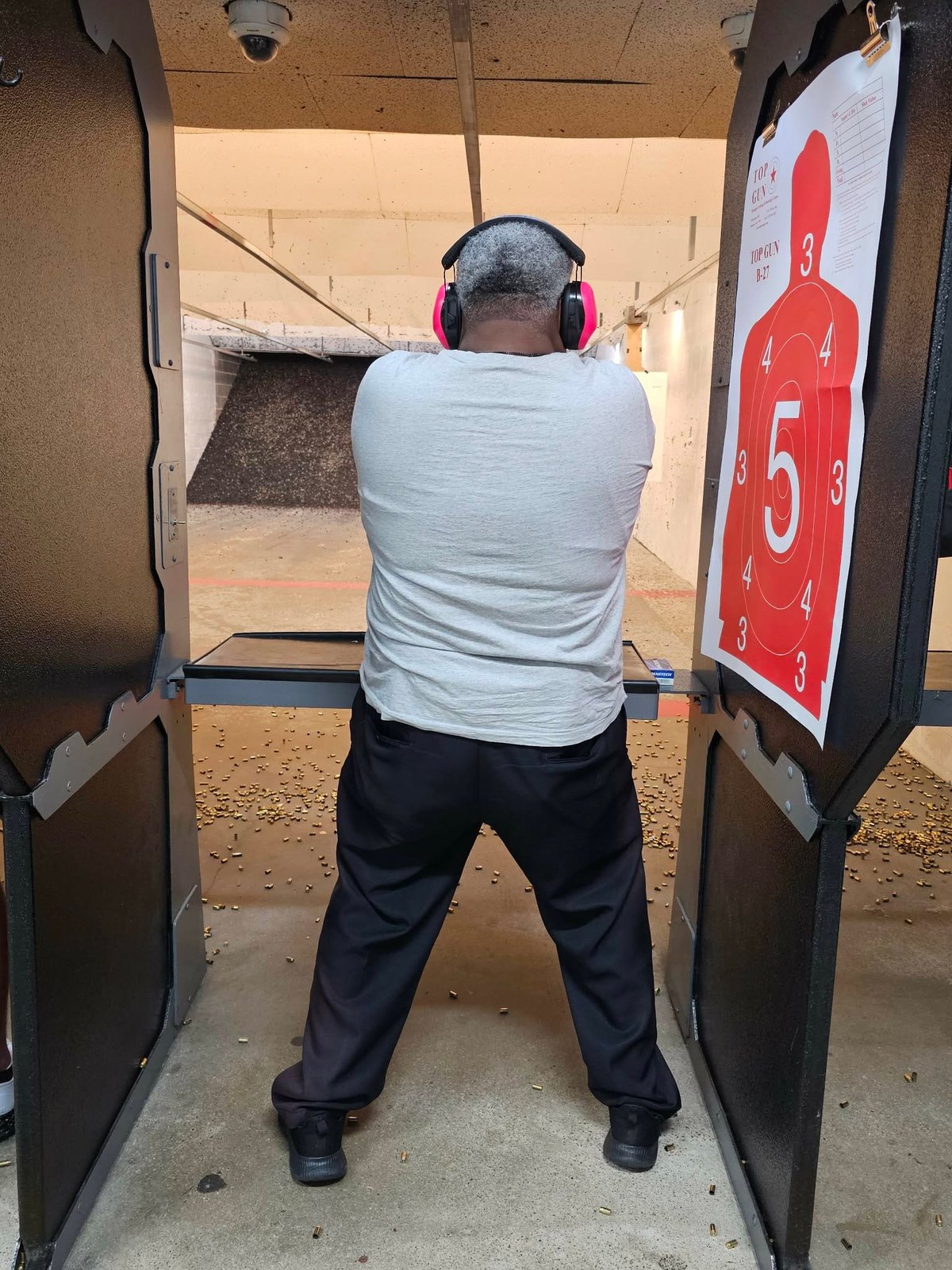 Person wearing headphones and white shirt at indoor shooting range, aiming at target with gun