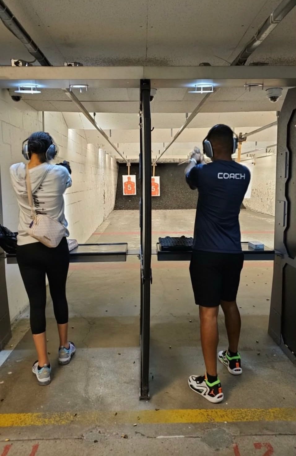 Two people at an indoor shooting range wearing headphones and facing target stalls with orange targets visible in the distance