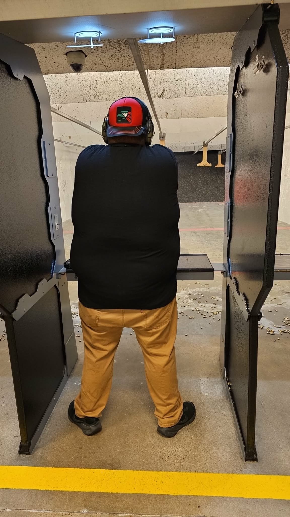 Person wearing red helmet and yellow pants at indoor shooting range, viewed from behind through open booth doors