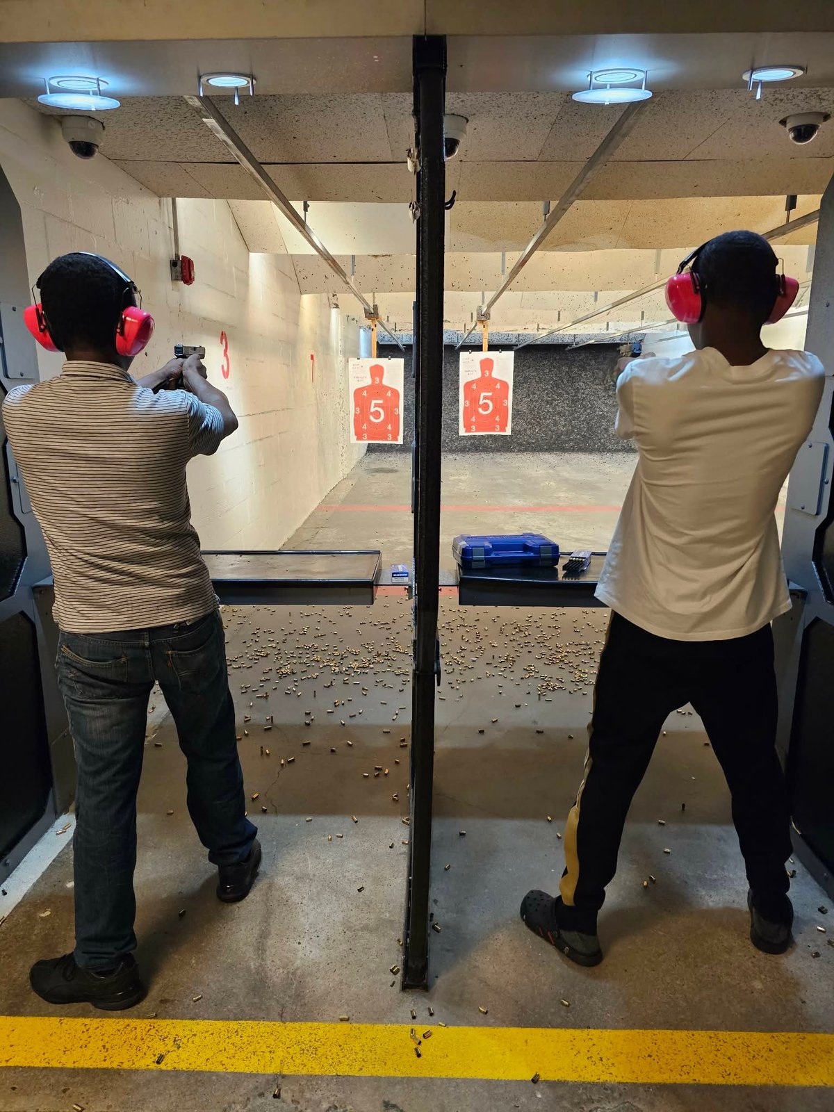 Two shooters at a gun range in separate booths wearing pink ear protection, aiming at numbered targets downrange