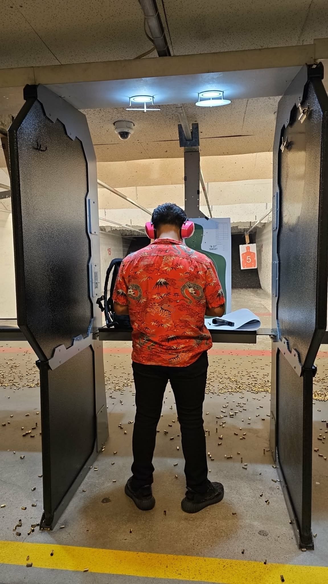 Person in red shirt standing at indoor shooting range between metal barriers, aiming at target downrange