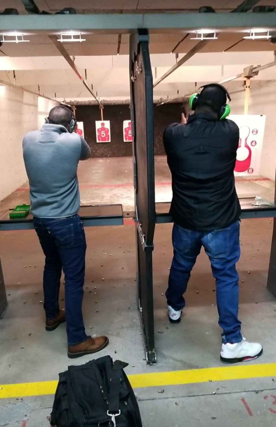 Two people wearing ear protection at an indoor shooting range, aiming at targets downrange