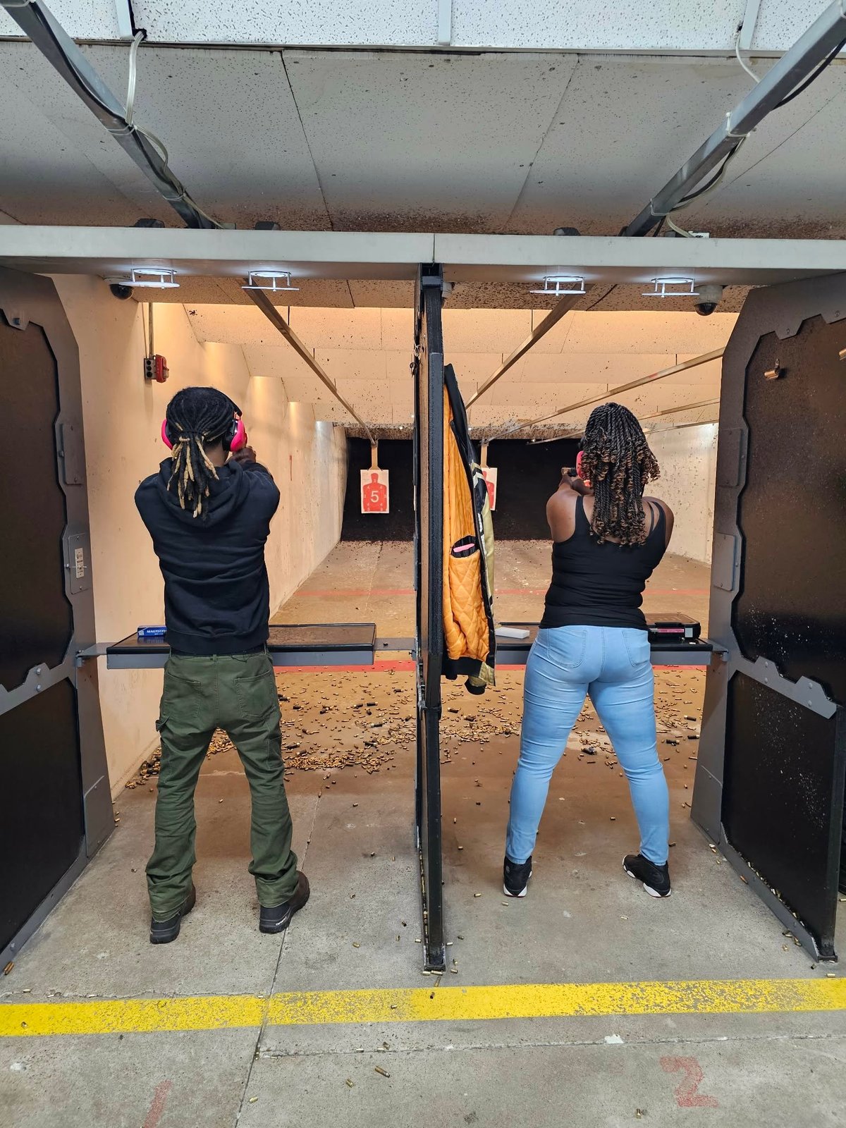 Two people at an indoor shooting range firing weapons at distant targets on a concrete floor