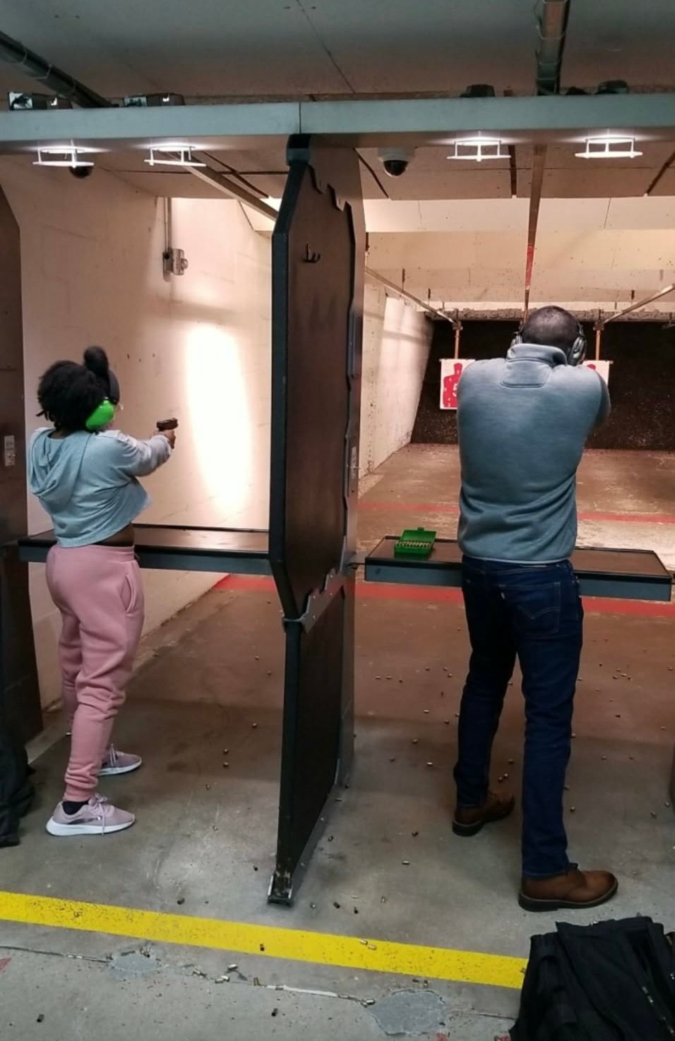 Two people at a shooting range, one on each side of a divider, aiming at targets with firearms