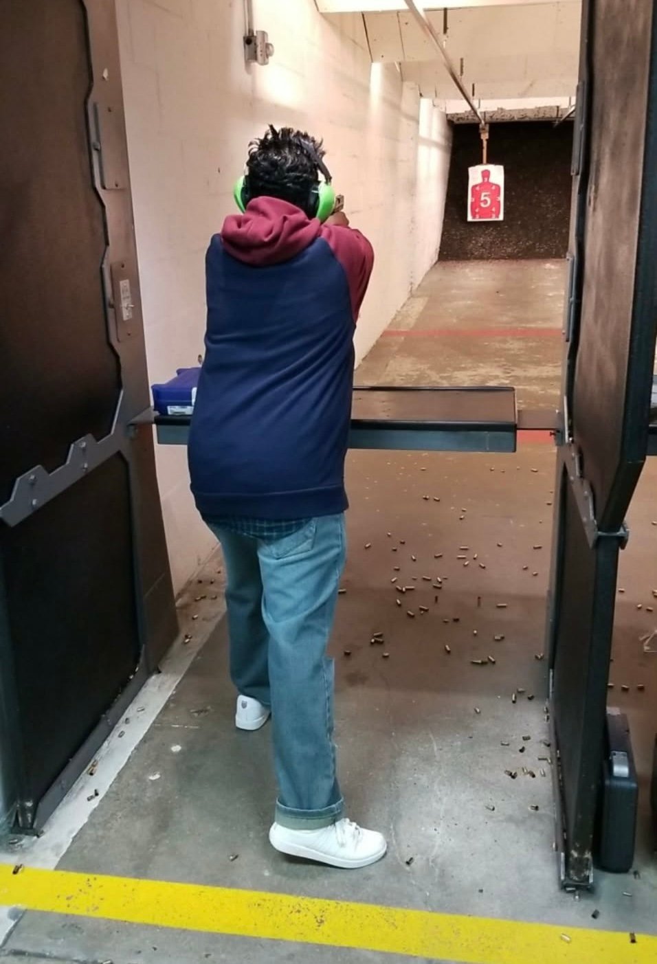 Person shooting at target in indoor range wearing blue and pink jacket facing downrange