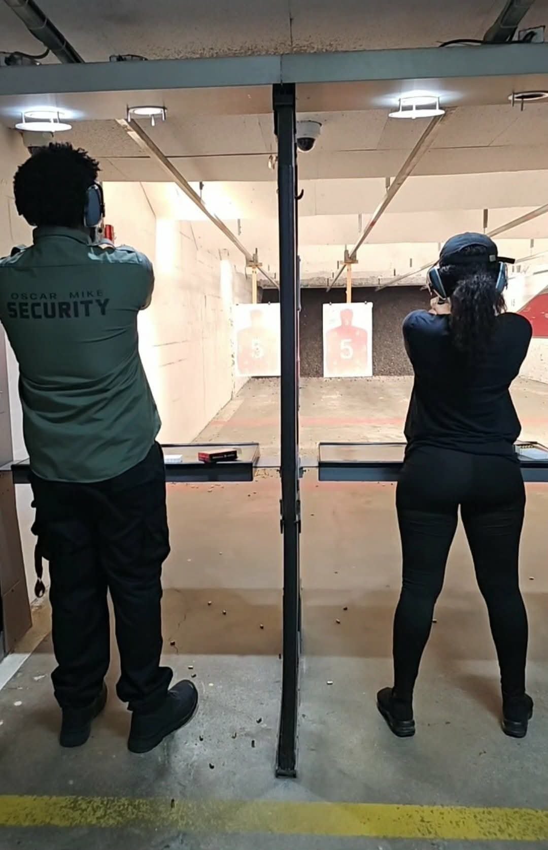 Two people at an indoor shooting range; security guard on left and person in cap on right aiming at targets downrange
