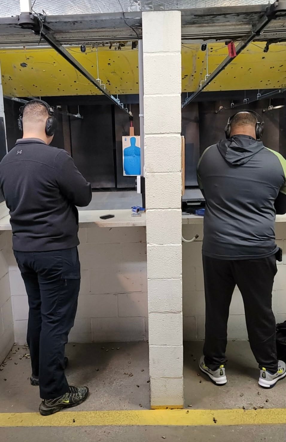 Two people wearing headphones practice shooting at an indoor gun range with target stands and yellow range dividers