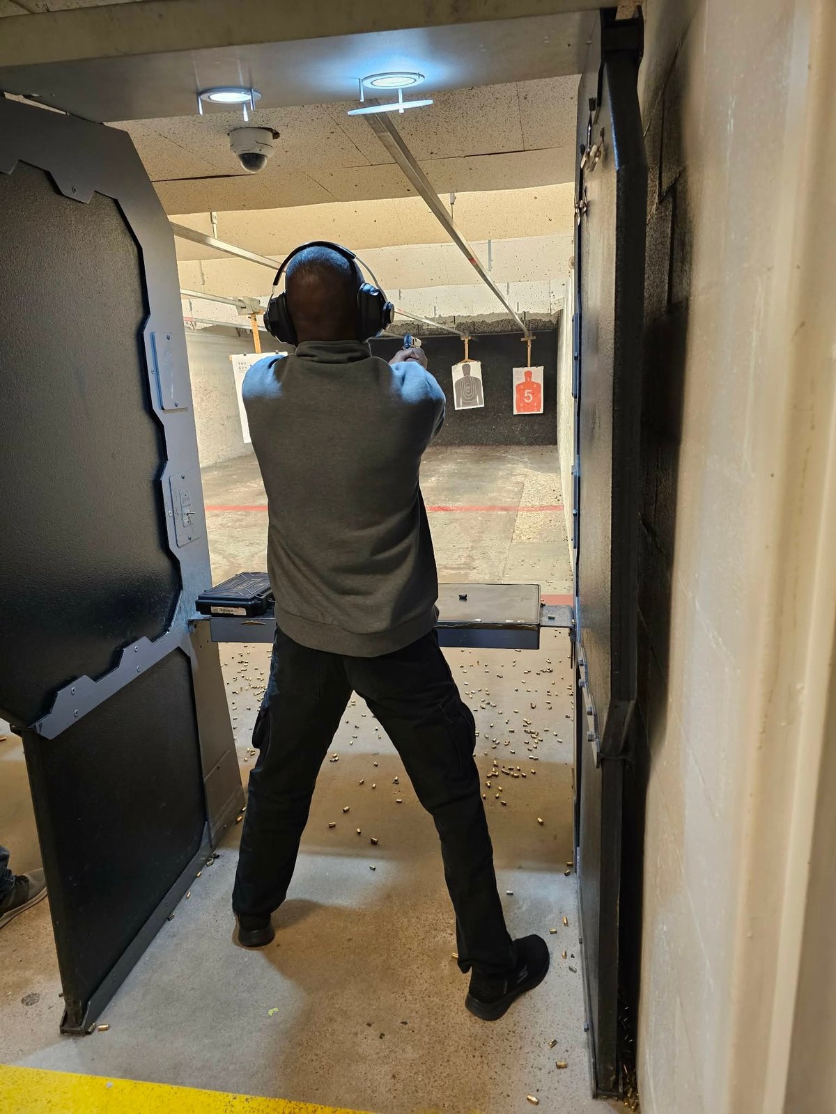 Person wearing headphones and protective gear aiming a firearm at targets in an indoor shooting range