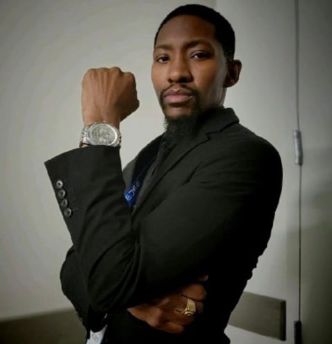 Man in black blazer posing with arm raised, displaying a large silver watch against a plain white wall background