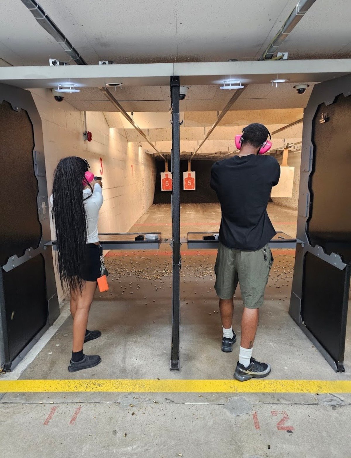 Two people wearing pink ear protection at an indoor shooting range, aiming at targets downrange