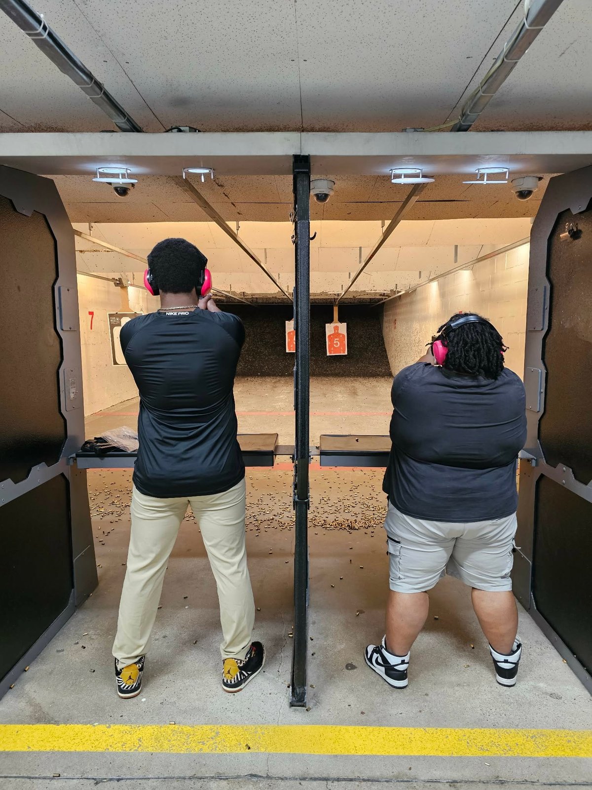 Two people wearing pink headphones and black shirts at an indoor shooting range aiming at targets downrange