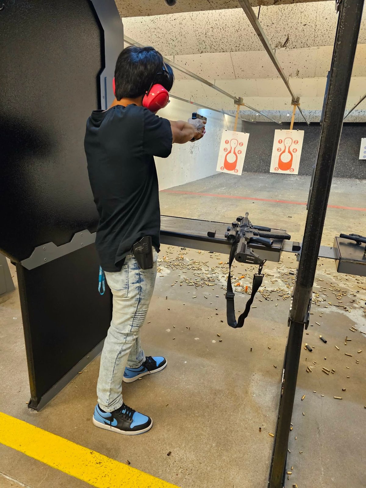 Person wearing red ear protection firing a rifle at a target range with paper targets visible downrange