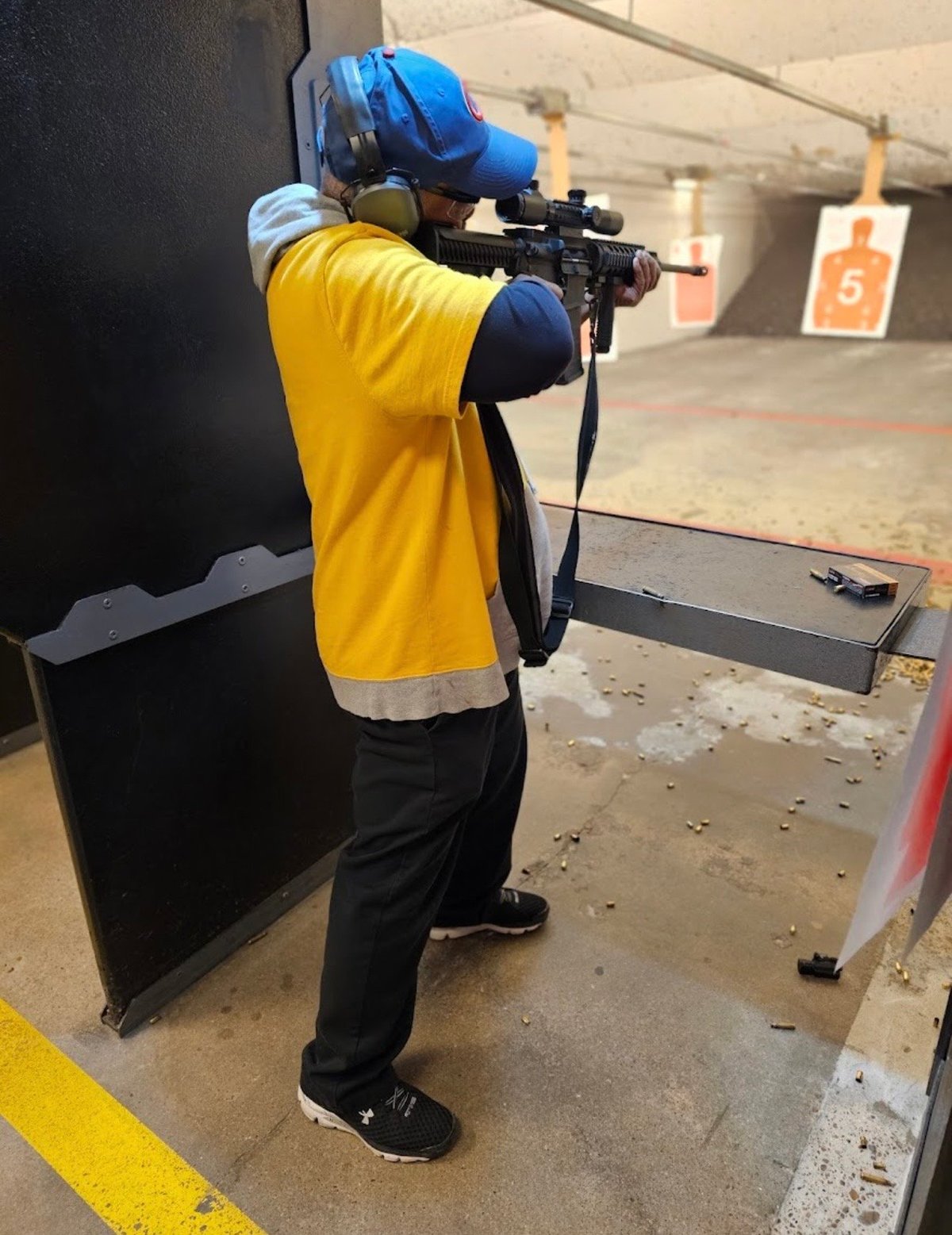 Shooter at indoor range wearing blue helmet and yellow shirt aiming rifle at target downrange