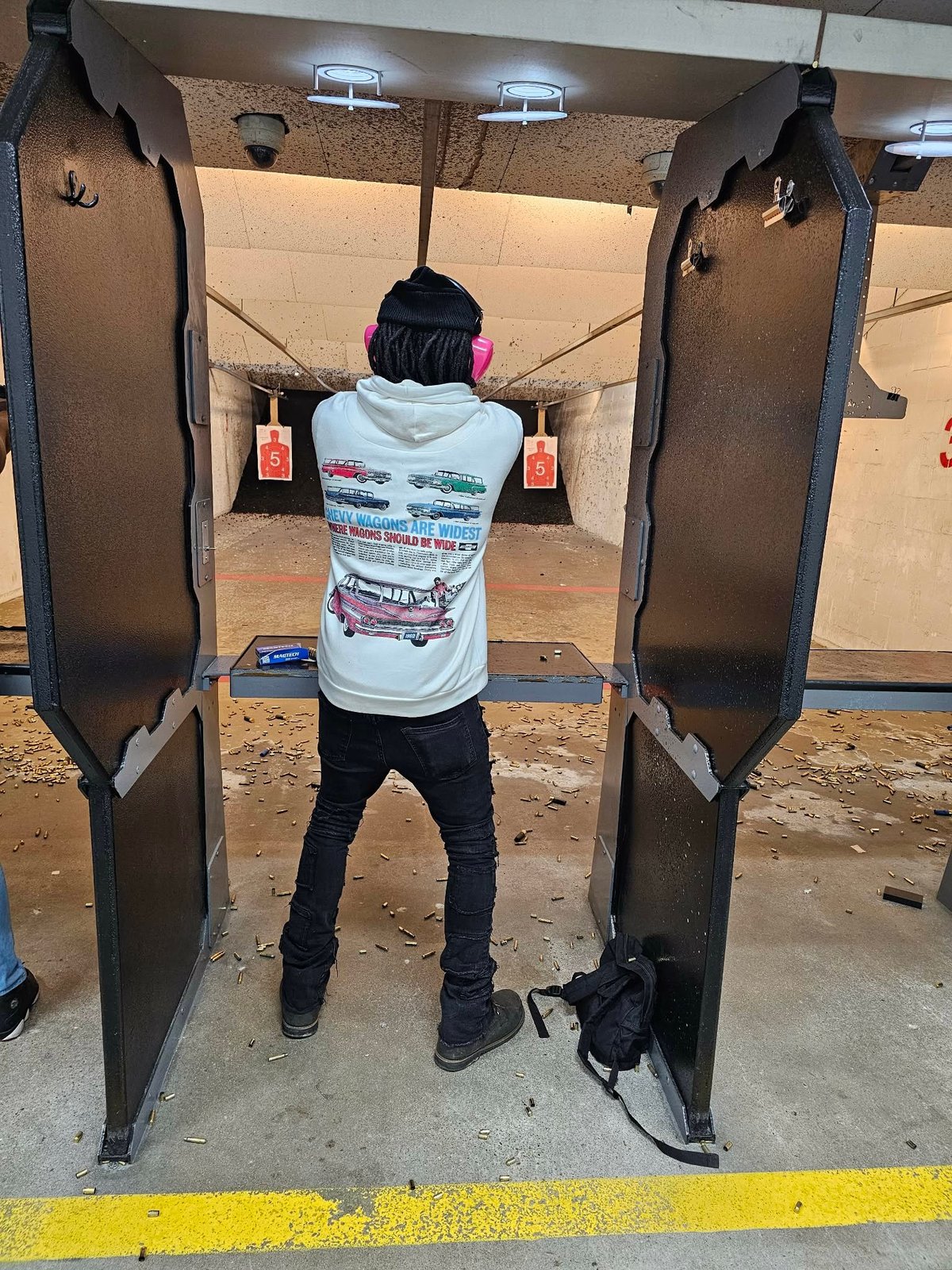 Person at indoor shooting range standing in firing booth facing targets, wearing hearing protection and holding firearm.